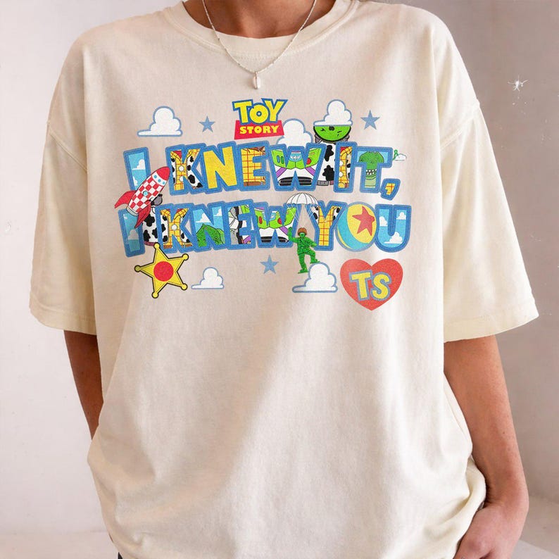 May include: A cream-colored short-sleeve t-shirt featuring a colorful Toy Story graphic. The design includes the text I KNEW IT, I KNEW YOU in blue letters filled with character patterns, a red rocket, a yellow star, clouds, and a red heart with TS.