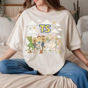 May include: A beige short-sleeve t-shirt featuring a graphic print of multiple cartoon characters in various outfits arranged in a row. Above the characters, the letters TS are printed in large blue and yellow text. The background includes clouds and stars.