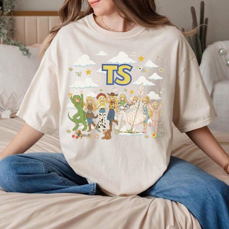 May include: A beige short-sleeve t-shirt featuring a graphic print of multiple cartoon characters in various outfits arranged in a row. Above the characters, the letters TS are printed in large blue and yellow text. The background includes clouds and stars.