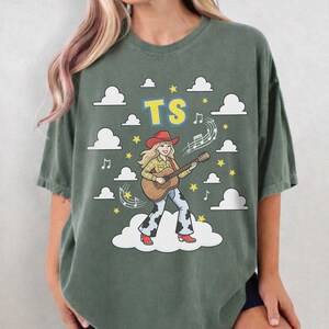 May include: A dark olive green t-shirt featuring a graphic of a blonde person wearing a red cowboy hat, yellow plaid shirt, and cow-print trousers while playing an acoustic guitar. The text TS appears above, surrounded by white clouds and yellow stars.