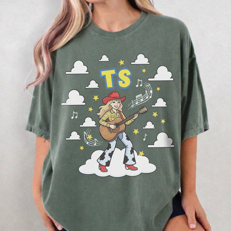 May include: A dark olive green t-shirt featuring a graphic of a blonde person wearing a red cowboy hat, yellow plaid shirt, and cow-print trousers while playing an acoustic guitar. The text TS appears above, surrounded by white clouds and yellow stars.