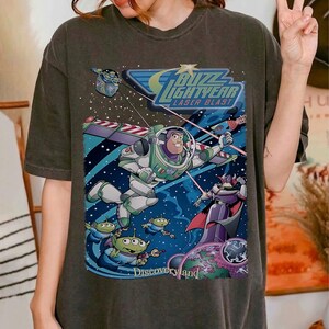 May include: Dark gray t-shirt featuring a colorful graphic from the movie Toy Story. The design includes Buzz Lightyear in a spacesuit, aliens, and the text "Buzz Lightyear Laser Blast" and "Discoveryland".