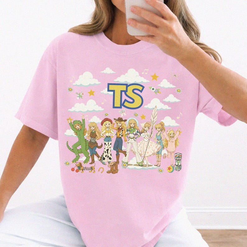 May include: A light pink t-shirt featuring a graphic print of multiple anime-style characters in various outfits, including a dinosaur costume and cowboy attire. The text TS is printed in large yellow letters above the characters, surrounded by clouds, stars, and musical notes.