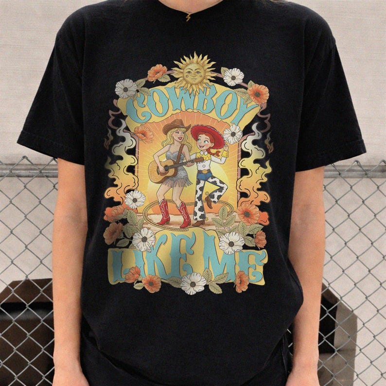 Puede incluir: Una camiseta negra de manga corta con un estampado gr&aacute;fico de dos figuras femeninas con atuendo de vaquera, una de ellas tocando una guitarra ac&uacute;stica. El dise&ntilde;o incluye el texto "COWBOY LIKE ME", un sol y detalles florales en naranja, amarillo y blanco.