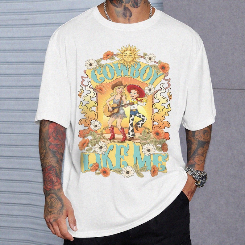 Puede incluir: Una camiseta blanca de manga corta con un estampado gr&aacute;fico de dos personajes de dibujos animados, uno tocando la guitarra, rodeados de flores y un sol. El texto "COWBOY LIKE ME" est&aacute; impreso en letras azules grandes sobre la ilustraci&oacute;n.