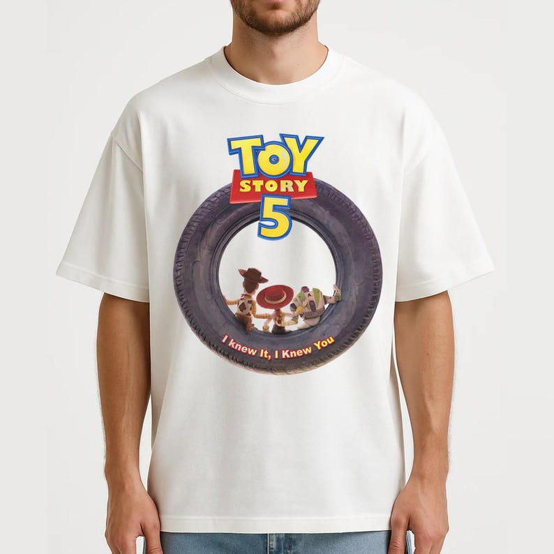 May include: A white short-sleeve t-shirt featuring a graphic of a black tire. Inside the tire are characters Woody, Jessie, and Buzz Lightyear. Above the tire is the text Toy Story 5, and below the tire is the text I knew it, I Knew You.