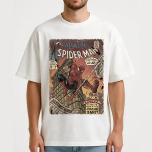 May include: White t-shirt featuring a vintage comic book cover of Spider-Man. The comic cover shows Spider-Man swinging from a building, with text that reads "The Amazing Spider-Man" and "Daily Bugle".