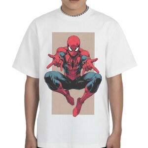 Pu&ograve; includere: Una t-shirt bianca a maniche corte con un'illustrazione grafica centrale del personaggio dei fumetti Spider-Man in posa accovacciata con le mani tese in avanti. Il personaggio indossa un costume rosso e blu con un emblema a ragno nero.