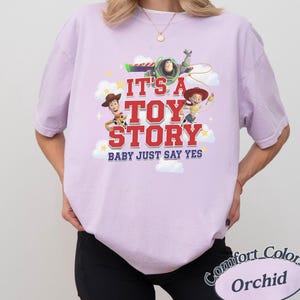 K&ouml;nnte beinhalten: Ein hellviolettes Kurzarm-T-Shirt mit einem Grafikdruck von Woody, Buzz Lightyear und Jessie aus Toy Story. Der Text lautet IT'S A TOY STORY BABY JUST SAY YES, umgeben von wei&szlig;en Wolken und gelben Sternen auf einem lavendelfarbenen Stoffhintergrund.