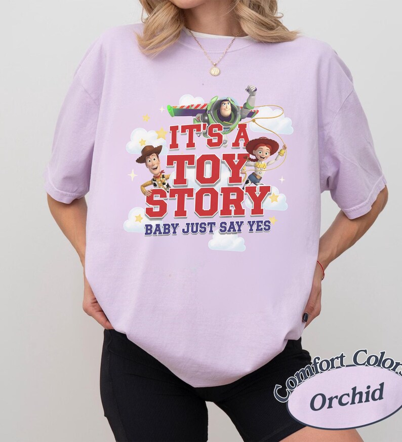 K&ouml;nnte beinhalten: Ein hellviolettes Kurzarm-T-Shirt mit einem Grafikdruck von Woody, Buzz Lightyear und Jessie aus Toy Story. Der Text lautet IT'S A TOY STORY BABY JUST SAY YES, umgeben von wei&szlig;en Wolken und gelben Sternen auf einem lavendelfarbenen Stoffhintergrund.
