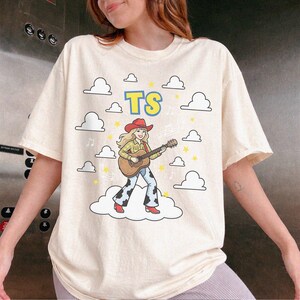 May include: A cream-coloured short-sleeve t-shirt featuring a graphic of a blonde figure in a red cowboy hat, yellow plaid shirt, and cow-print trousers playing an acoustic guitar. The design includes the letters TS, white clouds, and yellow stars.