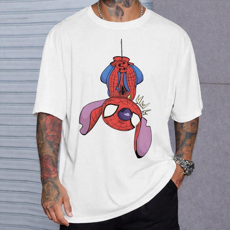 May include: A white short-sleeved t-shirt featuring a graphic print of a character resembling Stitch wearing a red and blue spider-themed costume, hanging upside down by a web line. The character has large ears and a mask with white eye lenses.