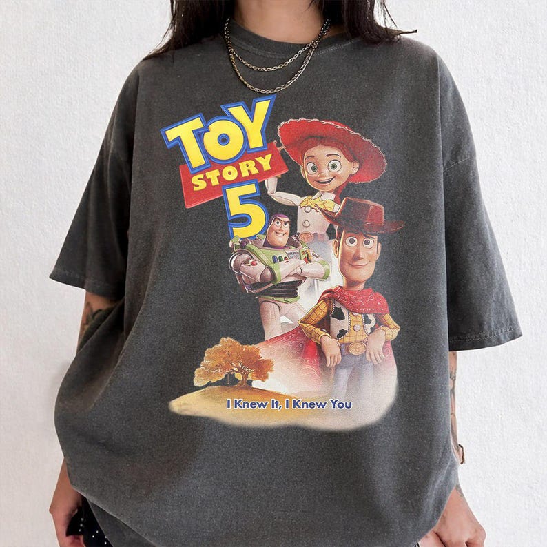 May include: A charcoal gray short-sleeve t-shirt featuring a graphic print of Toy Story 5 with characters Jessie, Buzz Lightyear, and Woody. The design includes the text Toy Story 5 and the phrase I Knew It, I Knew You above a small landscape.