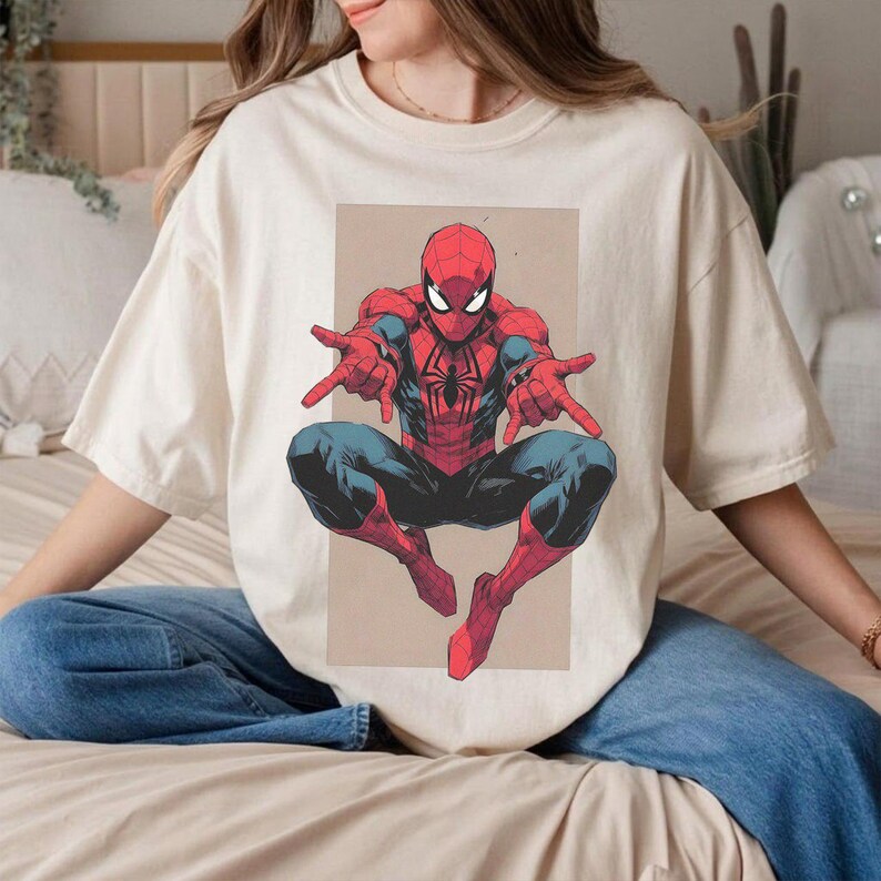 Pu&ograve; includere: Una t-shirt a maniche corte color crema con una stampa grafica del personaggio dei fumetti Spider-Man in posizione accovacciata. Il personaggio &egrave; raffigurato con un costume rosso e blu con un emblema a ragno nero sul petto e occhi bianchi.