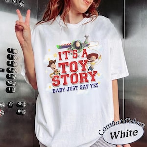 K&ouml;nnte beinhalten: Ein wei&szlig;es T-Shirt mit einem Grafikdruck von Woody, Buzz Lightyear und Jessie aus Toy Story. Der Text lautet IT'S A TOY STORY BABY JUST SAY YES in roter und blauer Schrift, umgeben von gelben Sternen und wei&szlig;en Wolken auf der Vorderseite.
