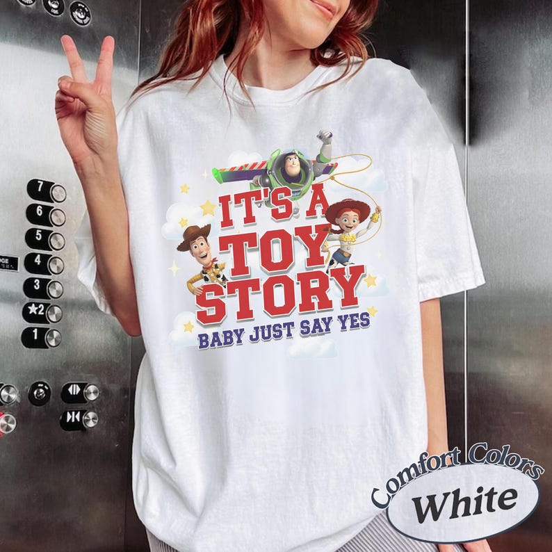 K&ouml;nnte beinhalten: Ein wei&szlig;es T-Shirt mit einem Grafikdruck von Woody, Buzz Lightyear und Jessie aus Toy Story. Der Text lautet IT'S A TOY STORY BABY JUST SAY YES in roter und blauer Schrift, umgeben von gelben Sternen und wei&szlig;en Wolken auf der Vorderseite.