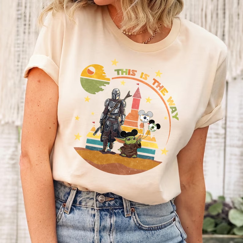 Puede incluir: Camiseta color crema con un dise&ntilde;o gr&aacute;fico. El dise&ntilde;o presenta el texto "THIS IS THE WAY", una figura mandaloriana, Grogu y globos con tem&aacute;tica de Mickey Mouse. El dise&ntilde;o est&aacute; sobre un fondo colorido con estrellas.