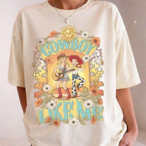 Puede incluir: Una camiseta de color crema con un estampado gr&aacute;fico de dos vaqueras de dibujos animados, una de ellas tocando una guitarra ac&uacute;stica, rodeadas de flores, un sol y llamas. El texto "COWBOY LIKE ME" est&aacute; impreso en letras grandes de color azul claro.
