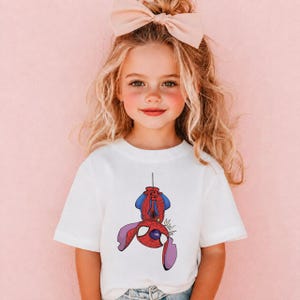 May include: A white short-sleeved t-shirt featuring a graphic print of a purple alien character wearing a red and blue superhero mask and suit, hanging upside down by a thin black line. The character has large ears and a stylized comic book design.