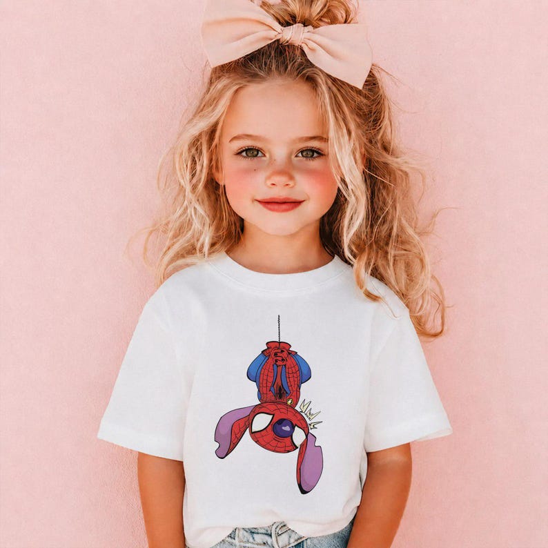 May include: A white short-sleeved t-shirt featuring a graphic print of a purple alien character wearing a red and blue superhero mask and suit, hanging upside down by a thin black line. The character has large ears and a stylized comic book design.