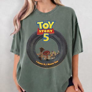 May include: A sage green short-sleeve t-shirt featuring a graphic of a black tire. Inside the tire are characters Woody, Jessie, and Buzz Lightyear. Above the tire is the text TOY STORY 5, and below the tire is the text I knew it, I knew You.