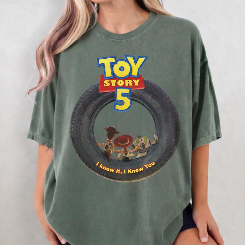 May include: A sage green short-sleeve t-shirt featuring a graphic of a black tire. Inside the tire are characters Woody, Jessie, and Buzz Lightyear. Above the tire is the text TOY STORY 5, and below the tire is the text I knew it, I knew You.