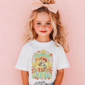 Puede incluir: Una camiseta blanca de manga corta con un estampado gr&aacute;fico de una vaquera tocando la guitarra y un vaquero bailando. El dise&ntilde;o incluye el texto COWBOY LIKE ME, un sol y elementos florales en tonos amarillos, naranjas, azules y blancos.