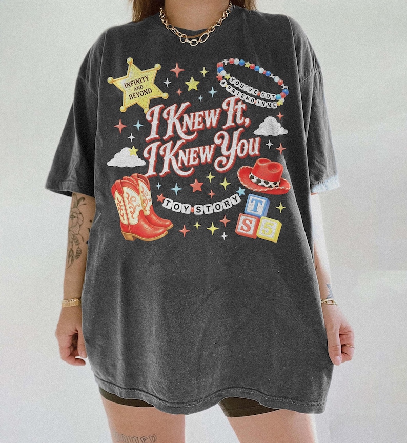May include: A charcoal grey short-sleeve t-shirt featuring a graphic print with the text I KNEW IT, I KNEW YOU, TOY STORY, INFINITY AND BEYOND, and YOU'VE GOT A FRIEND IN ME. The design includes a yellow star, cowboy boots, a hat, and blocks.