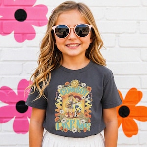 Puede incluir: Una camiseta de manga corta gris carb&oacute;n con un estampado gr&aacute;fico que incluye el texto COWBOY LIKE ME. El dise&ntilde;o presenta dos figuras ilustradas, un sol y elementos florales en tonos amarillos, naranjas y blancos en la parte delantera.