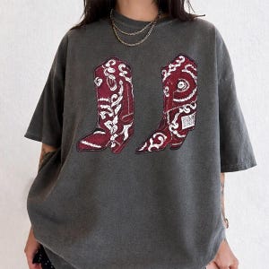 K&ouml;nnte beinhalten: Ein anthrazitfarbenes Kurzarm-T-Shirt mit einem Grafikdruck von zwei kastanienbraunen Cowboystiefeln mit komplizierten wei&szlig;en Paisley- und Blumenmustern. Das T-Shirt hat einen Rundhalsausschnitt und eine lockere, &uuml;bergro&szlig;e Passform aus Vintage-Stoff.