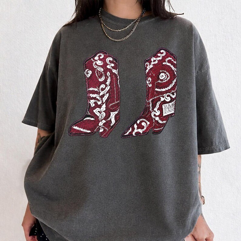 K&ouml;nnte beinhalten: Ein anthrazitfarbenes Kurzarm-T-Shirt mit einem Grafikdruck von zwei kastanienbraunen Cowboystiefeln mit komplizierten wei&szlig;en Paisley- und Blumenmustern. Das T-Shirt hat einen Rundhalsausschnitt und eine lockere, &uuml;bergro&szlig;e Passform aus Vintage-Stoff.
