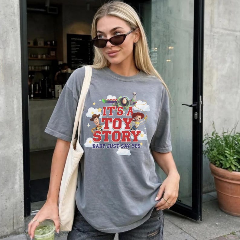 K&ouml;nnte beinhalten: Ein anthrazitfarbenes Kurzarm-T-Shirt mit einem Grafikdruck von Woody, Buzz Lightyear und Jessie aus Toy Story. Der Text lautet IT'S A TOY STORY BABY JUST SAY YES, umgeben von wei&szlig;en Wolken und gelben Sternen auf der Vorderseite des Shirts.