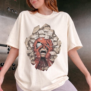 May include: A cream-colored short-sleeve t-shirt featuring a graphic print of a red masked character with hands pressed against their face in distress, surrounded by a cluster of floating, paper-like geometric shapes covered in handwritten numbers.