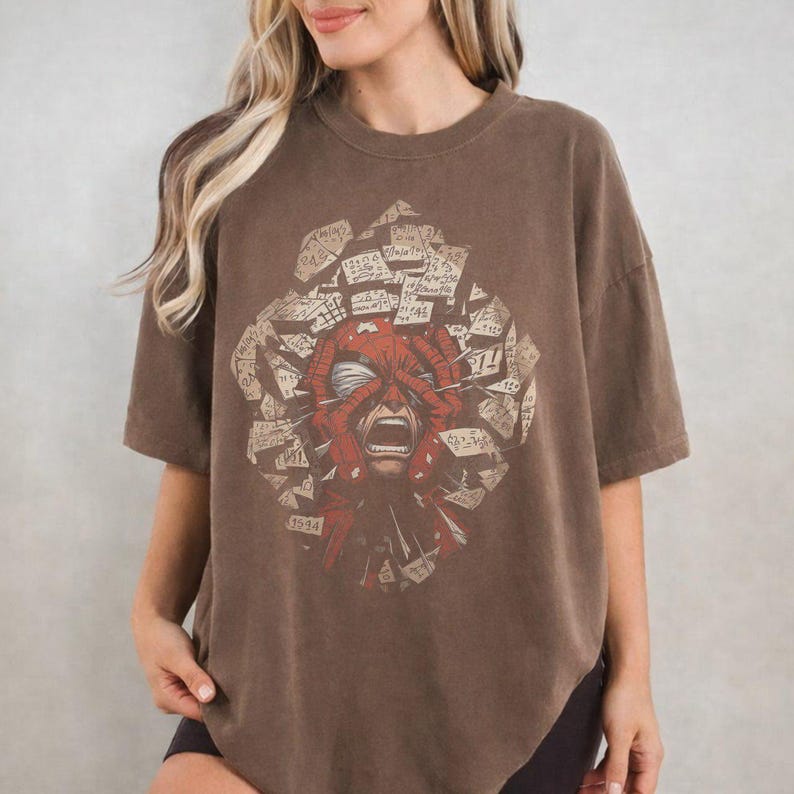 May include: A brown short-sleeve t-shirt featuring a central graphic of a red masked figure with hands covering its face, surrounded by a collage of scattered rectangular paper fragments containing handwritten text and various numerical figures.