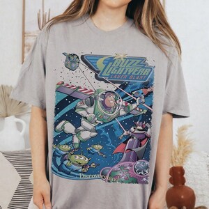 May include: A gray t-shirt featuring a colorful graphic of Buzz Lightyear from Toy Story in a space scene. The design includes the text "Buzz Lightyear Laser Blast" and "Discoveryland".