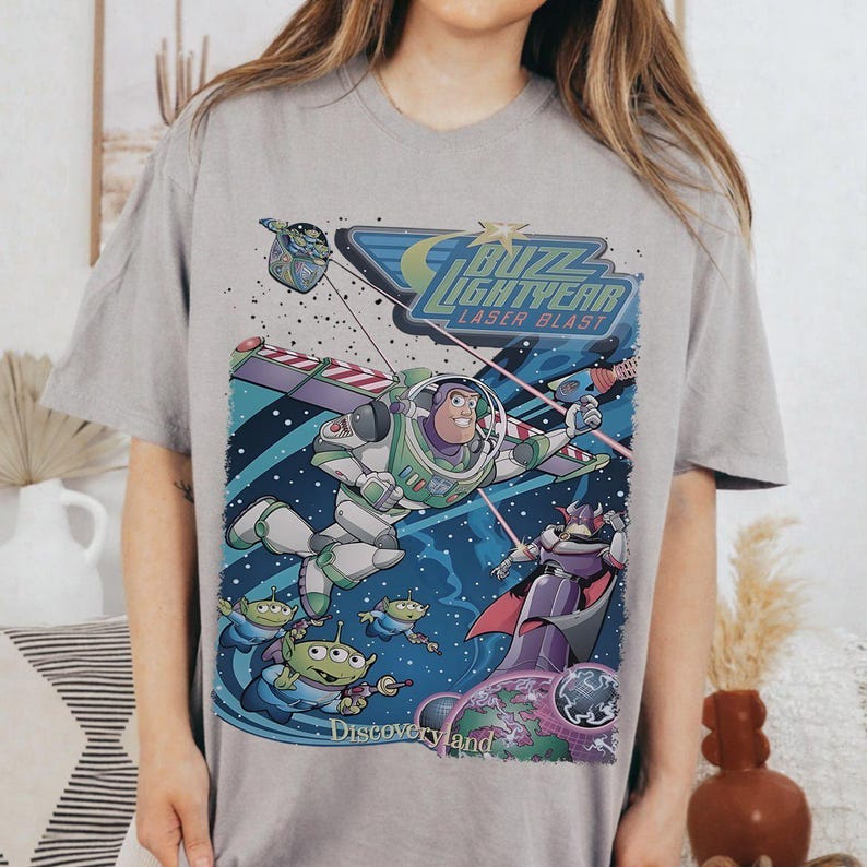 May include: A gray t-shirt featuring a colorful graphic of Buzz Lightyear from Toy Story in a space scene. The design includes the text "Buzz Lightyear Laser Blast" and "Discoveryland".