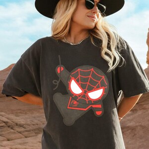 May include: A charcoal grey short-sleeve t-shirt featuring a stylised graphic of a cartoon character with a red spiderweb pattern on the head, large white eyes, and a small red spider icon on the chest area, printed in a bold, minimalist design.
