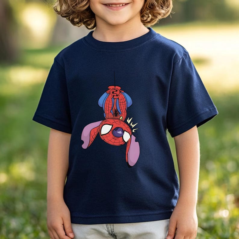 May include: A navy blue short-sleeve t-shirt featuring a graphic print of a character resembling Stitch dressed in a red and blue Spider-Man costume, hanging upside down by a web line with large white eyes and purple ears visible.