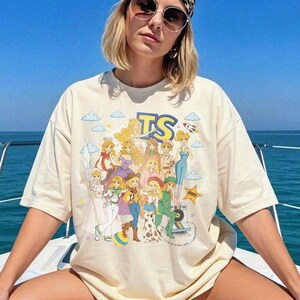 May include: A cream-colored short-sleeve t-shirt featuring a large graphic print of multiple cartoon characters in various outfits. The text TS is printed in large blue and yellow letters at the top, surrounded by small clouds, stars, and a rocket.