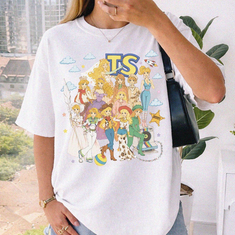 May include: A white short-sleeve t-shirt featuring a colorful graphic print of various animated characters in a retro style. The design includes the text TS at the top, a rocket, clouds, a sheriff star, and a vintage camera. The shirt has a crew neckline.
