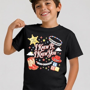 May include: A black short-sleeved t-shirt featuring a graphic print with the text I KNEW IT, I KNEW YOU, TOY STORY, INFINITY AND BEYOND, and YOU'VE GOT A FRIEND IN ME. The design includes a yellow sheriff star, red cowboy boots, a red hat, and blocks.