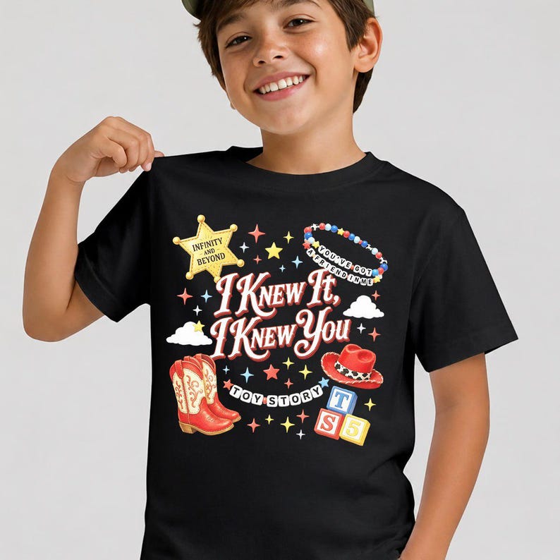 May include: A black short-sleeved t-shirt featuring a graphic print with the text I KNEW IT, I KNEW YOU, TOY STORY, INFINITY AND BEYOND, and YOU'VE GOT A FRIEND IN ME. The design includes a yellow sheriff star, red cowboy boots, a red hat, and blocks.