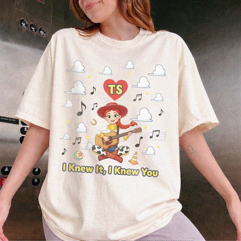 May include: A cream-colored t-shirt featuring a graphic of a cowgirl character playing an acoustic guitar. The design includes a red heart with the letters TS, musical notes, clouds, a ball, and a traffic cone, with the text I Knew It, I Knew You.