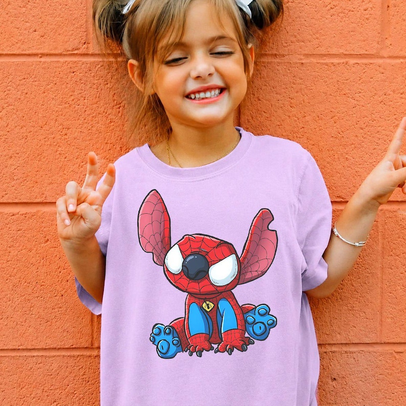 May include: A light purple short-sleeved t-shirt featuring a graphic print of a blue and red character with large ears and a spider-web pattern on its head, resembling a superhero mask, with blue accents on the feet and a small spider emblem.
