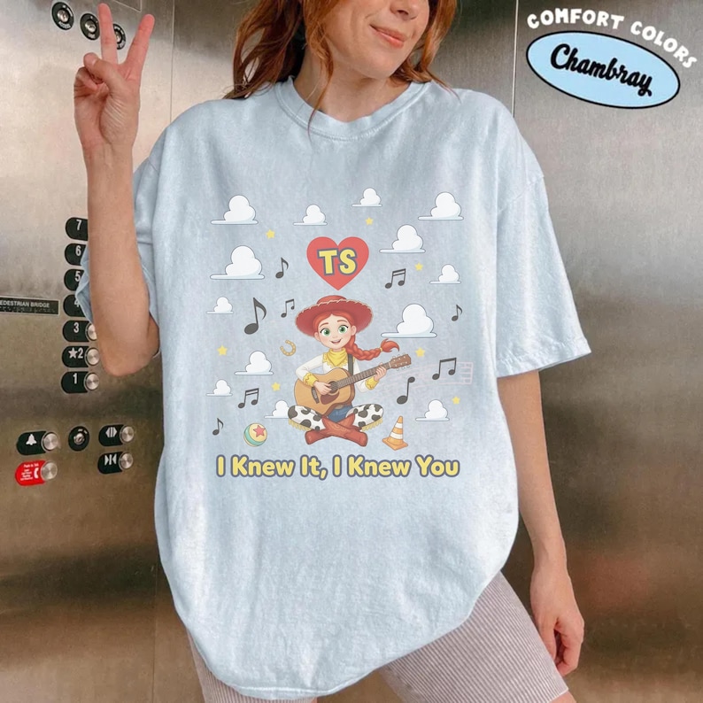 May include: A light blue t-shirt featuring a graphic of a cartoon cowgirl playing an acoustic guitar. The design includes a red heart with the letters TS, musical notes, clouds, a ball, and a traffic cone. The text reads I Knew It, I Knew You.