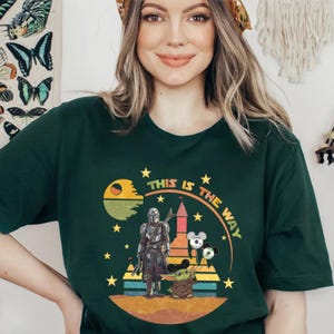 Puede incluir: Camiseta verde oscuro con un dise&ntilde;o gr&aacute;fico que dice "THIS IS THE WAY". El dise&ntilde;o incluye un personaje, un personaje beb&eacute; y un planeta. El dise&ntilde;o tambi&eacute;n incluye estrellas y un castillo.