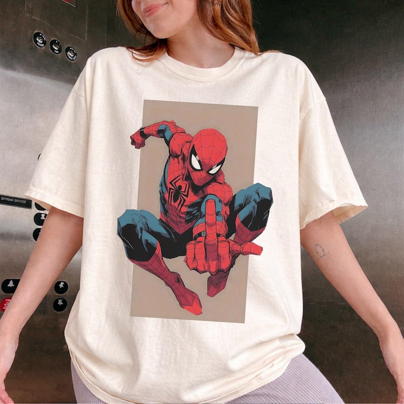 Puede incluir: Una camiseta de manga corta de color crema con un estampado gr&aacute;fico del personaje de c&oacute;mic Spider-Man en pose agachada. El personaje aparece con un traje rojo y azul con un emblema de ara&ntilde;a negra en el pecho, sobre un fondo rectangular de color tostado.