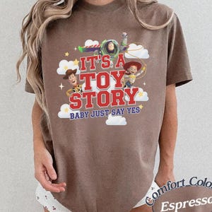 K&ouml;nnte beinhalten: Ein braunes Kurzarm-T-Shirt mit einem Grafikdruck von Woody, Buzz Lightyear und Jessie aus Toy Story. Der Text lautet IT'S A TOY STORY BABY JUST SAY YES, umgeben von wei&szlig;en Wolken und gelben Sternen auf der Vorderseite des Kleidungsst&uuml;cks.