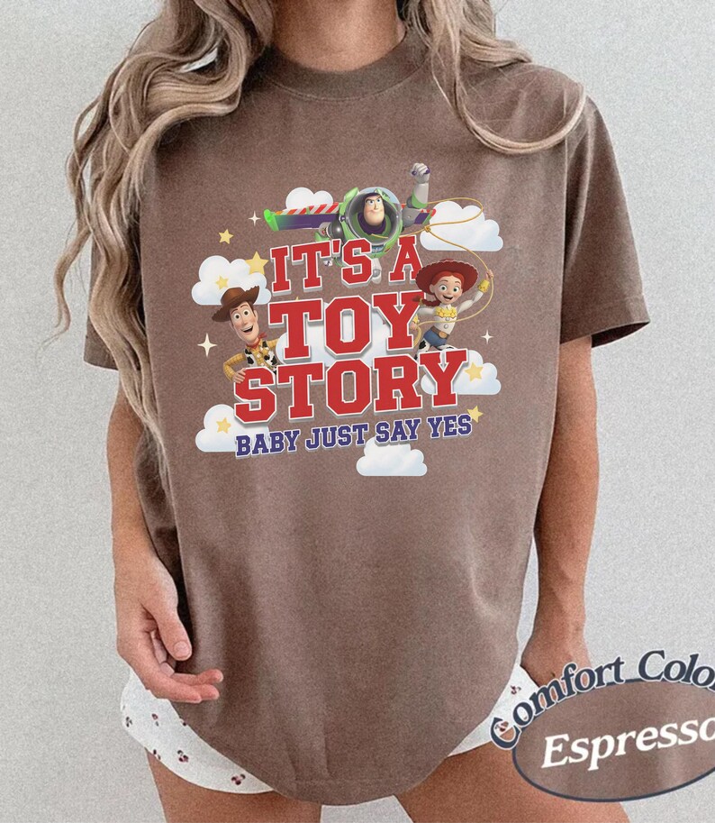 K&ouml;nnte beinhalten: Ein braunes Kurzarm-T-Shirt mit einem Grafikdruck von Woody, Buzz Lightyear und Jessie aus Toy Story. Der Text lautet IT'S A TOY STORY BABY JUST SAY YES, umgeben von wei&szlig;en Wolken und gelben Sternen auf der Vorderseite des Kleidungsst&uuml;cks.