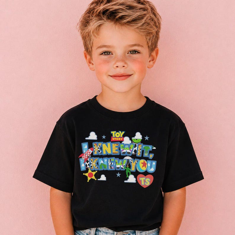 Pode incluir: Uma t-shirt preta de manga curta com um gr&aacute;fico colorido do Toy Story. O design inclui o texto 'I KNEW IT, I KNEW YOU' com padr&otilde;es de personagens, um foguet&atilde;o, uma estrela amarela, nuvens brancas e um cora&ccedil;&atilde;o vermelho com 'TS'.